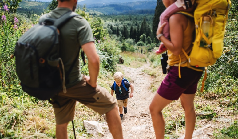 A family with two adults and two kids enjoys hiking on a forest trail. The child in the middle wears a blue backpack, while the adult on the right carries the other child.