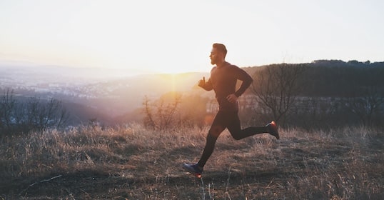 As morning light kisses the grassy field, a person, seemingly born to run, glides effortlessly through nature's embrace. With hills and trees painting the horizon, each step syncs with the rhythm of their internal podcast of thoughts and dreams.