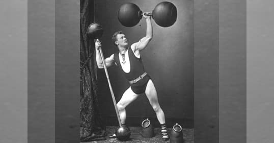 A vintage black and white photo captures the essence of Physical Culture, featuring a strongman in a singlet and striped socks, confidently posing with large, round weights.