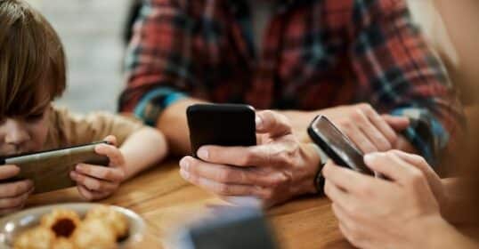 A family gathers around the table, each member engrossed in their smartphones as a podcast plays softly in the background. A bowl of snacks sits invitingly, offering a hint of intentional connection amidst the digital focus.