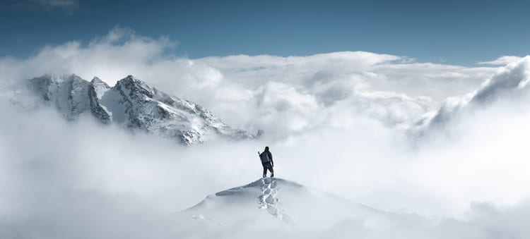 A high-agency individual stands atop a snow-covered mountain peak, surrounded by clouds, with a majestic mountain range in the background, showcasing the characteristics of determination and resilience.