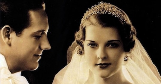 A black-and-white vintage wedding photo captures a historic moment of marriage as the groom gazes fondly at the bride, who faces the camera with a beaded tiara and veil.