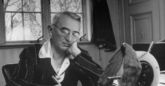 An elderly man in a suit writes at his desk, head resting on his hand, reflecting perhaps on the 