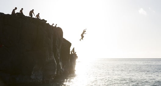 People are gathered on a rocky cliff by the sea, with one person mid-air jumping into the water.