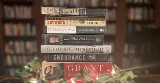 A stack of history books, perfect as a thoughtful Christmas gift, is surrounded by holly and lit with string lights, set against a background of blurred bookshelves.
