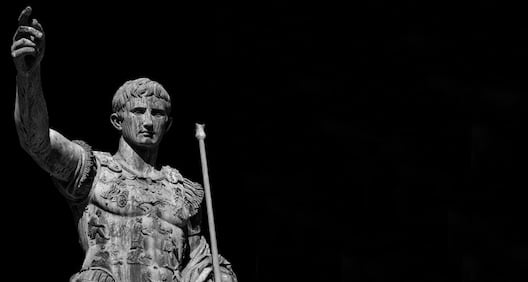 Black and white image of a Roman statue depicting a man in armor holding a staff, with one arm raised against a dark background, as if ready to share heroic tales on an ancient podcast.