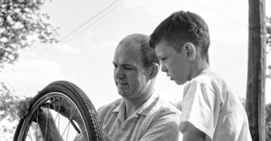 A man and a boy are repairing a bicycle tire outside. Demonstrating the skills of successful fathers, the man carefully explains each step while the boy watches closely, eager to learn.