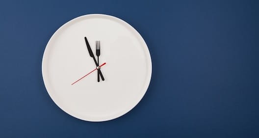 A clock resembling a white plate has black fork and knife hands, and a red second hand, set against a dark blue background.