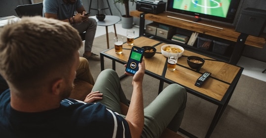 A person lounges on the couch, smartphone in hand with a betting app open, catching the soccer game on TV. Snacks and drinks adorn the coffee table as Episode 1044 of their favorite podcast plays softly in the background for some audio content multitasking.