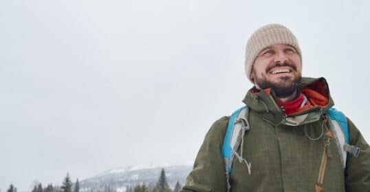 A person in a green jacket and beige beanie is hiking through a winter wonderland, with trees and a majestic mountain in the background.