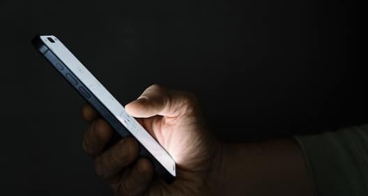 Close-up of a hand holding a smartphone in a dark setting, with the screen glowing and the user interacting with it.