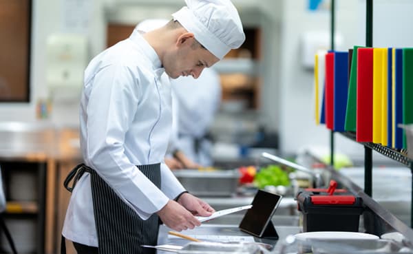 In a bustling commercial kitchen, a chef in a crisp white uniform examines a document—an embodiment of his secret to a productive and organized life. Nearby, colorful cutting boards rest neatly on a rack, exemplifying the harmony of mise en place.