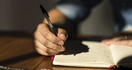 A person writes with a pen in a notebook on a wooden table, with sunlight illuminating the scene.