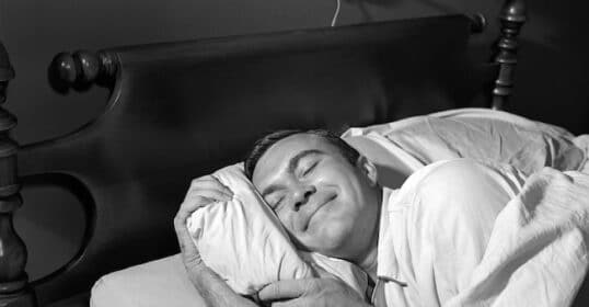 A man in pajamas lies in bed, smiling contentedly while hugging a pillow with both arms, showing how simple habits can help you improve your sleep.