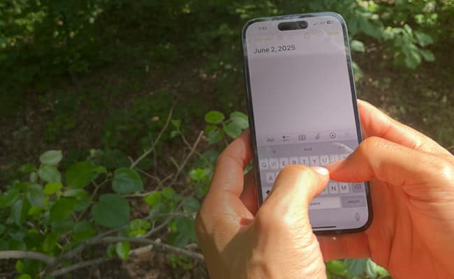 A person types "June 2, 2025" on a smartphone notes app while standing outdoors near green plants, embracing digital journaling to replace scrolling with mindful reflection.