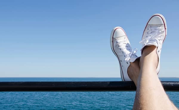 A person with crossed legs wearing white men's summer footwear rests their feet on a railing, overlooking a calm ocean under a clear blue sky.