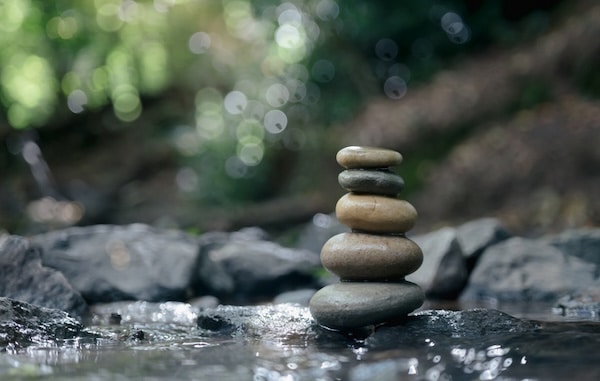 Five smooth stones stacked in a vertical pile on wet rocks beside a shallow stream, with blurred greenery in the background.