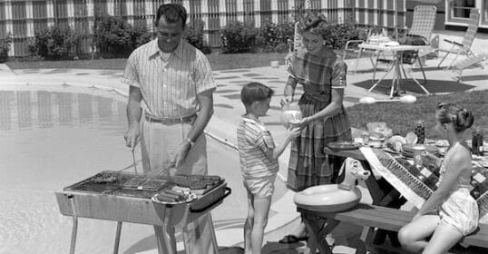 A man showcases suburban manhood as he grills by the pool, while a woman serves a boy and a girl sits at a picnic table surrounded by food and pool toys.