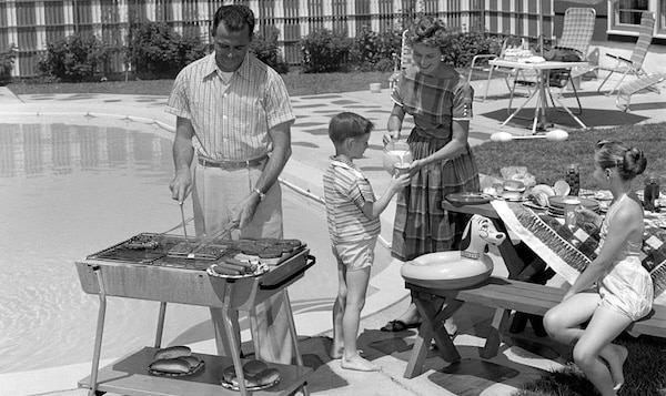 A man showcases suburban manhood as he grills by the pool, while a woman serves a boy and a girl sits at a picnic table surrounded by food and pool toys.