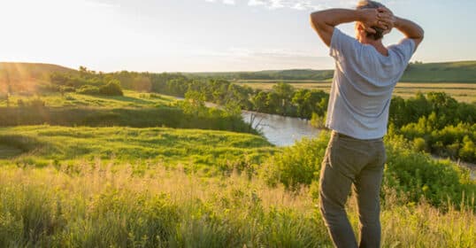 A person stands in a grassy field with hands on their head, reflecting during the summer solstice, looking out over a river and green landscape under a partly cloudy sky at sunset.