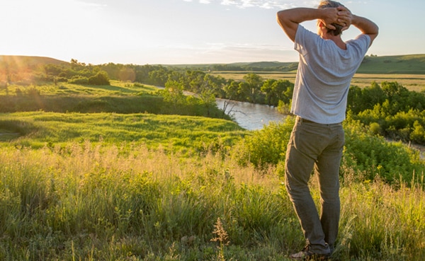 A person stands in a grassy field with hands on their head, reflecting during the summer solstice, looking out over a river and green landscape under a partly cloudy sky at sunset.