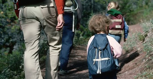 Adults and children wearing backpacks are walking together on a dirt trail through a wooded area, enjoying a family adventure and discovering the joys of backpacking with kids.