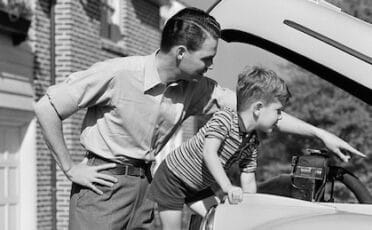 A man and a young boy look under the hood of a car; dressed in classic men's style, the duo inspects the engine as the boy points at something while the man observes.
