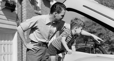 A man and a young boy look under the hood of a car; dressed in classic men's style, the duo inspects the engine as the boy points at something while the man observes.