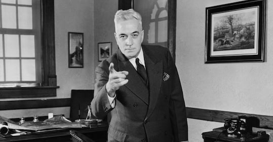 A man in a suit stands in an office, confidently pointing toward the camera, inspiring you to start executing plans, with a desk, window, and framed artwork visible in the background.