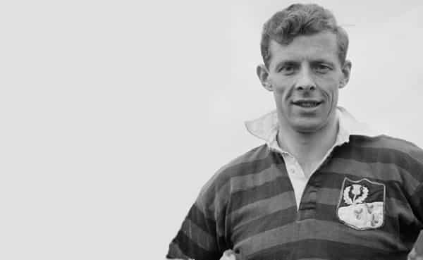 A man wearing a striped rugby shirt with an emblem stands outdoors, looking at the camera, against a plain light background—a classic look in men's fashion.