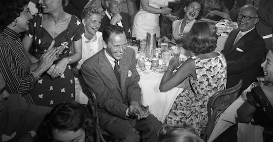 A group of well-dressed people sit and stand around a table, smiling and laughing in a lively social gathering—perhaps swapping stories about Frank Sinatra or sharing a clever tipping guide.