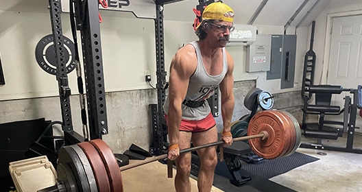 A man wearing a yellow headband and red shorts is focused on his fitness as he lifts a heavy barbell in a home gym equipped with weightlifting equipment.