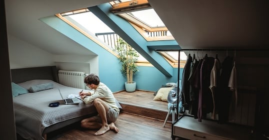 Person sitting on the floor of a modern attic bedroom, using a laptop on the bed near large skylight windows with clothes hanging on a rack nearby—an ideal spot to enjoy ultimate morning routine practices.
