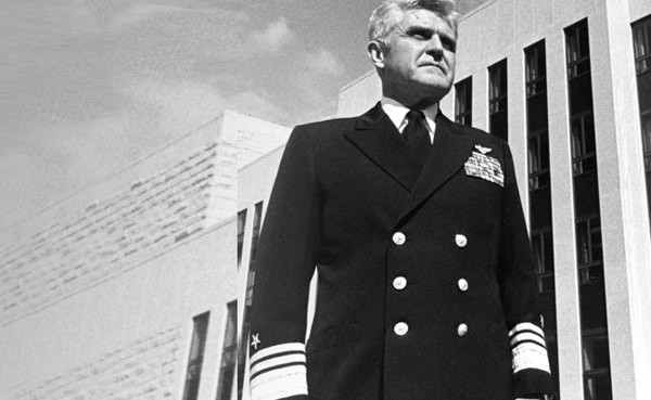 A man in a formal military uniform with medals stands outside a modern building, embodying the Stockdale Paradox as he looks into the distance, reflecting on tough times with unwavering resolve.