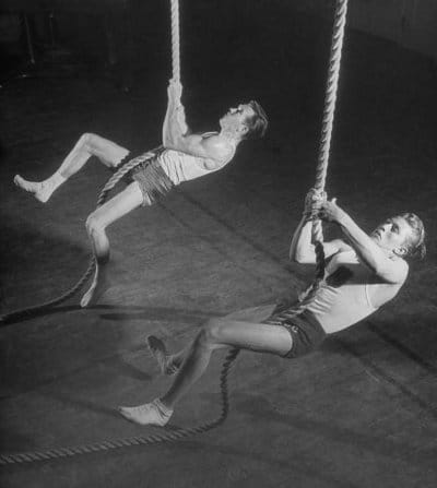 Vintage young men climbing ropes in a gym.