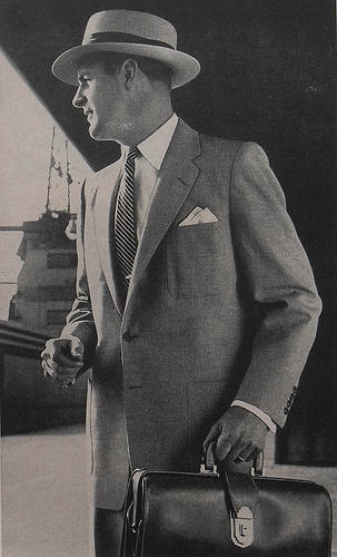 Mid 1900s businessman wearing suit and hat holding briefcase in hand.