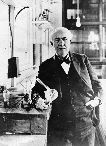 Vintage Thomas Edison standing in workshop and holding light bulb.