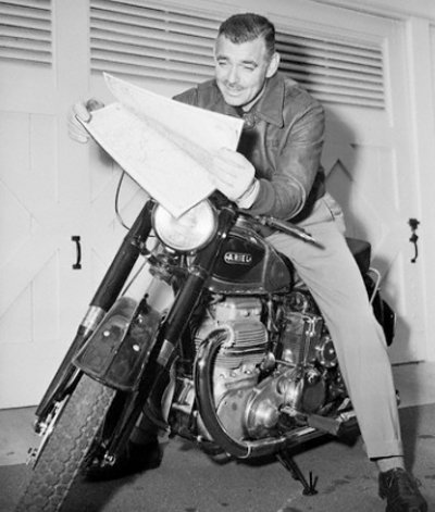 Clark Gable reading newspaper while riding on motorcycle. 