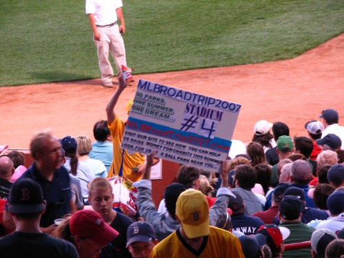 Man carrying MLB road trip board in crowded stadium.