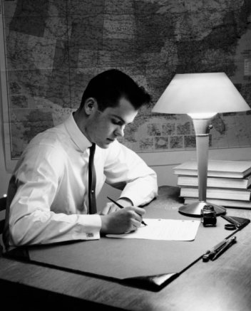 Vintage young man studying at desk in lamp light and a map on wall. 