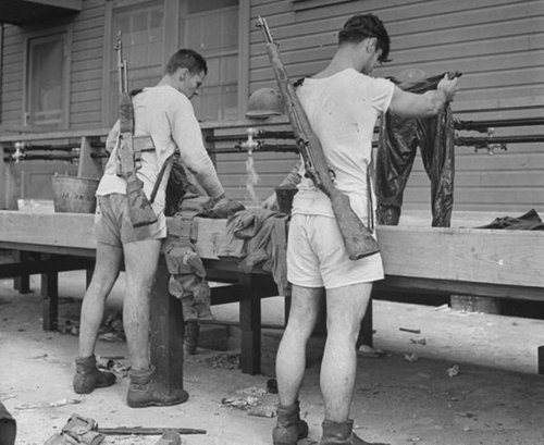 Military men in shorts washing clothes while wearing guns. 