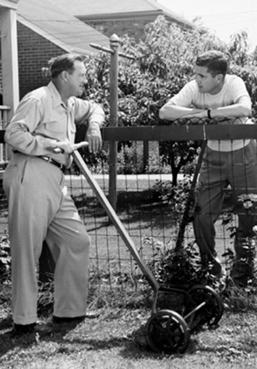 Vintage neighbors talking over fence mowing lawns. 
