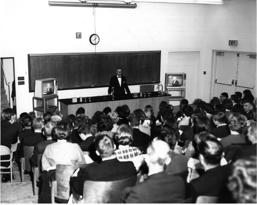 Vintage college class professor lecturing at front of classroom.