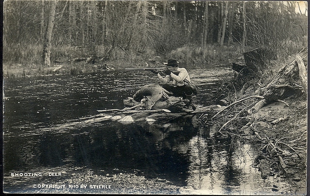 Vintage hunter aiming rifle on riverbank. 