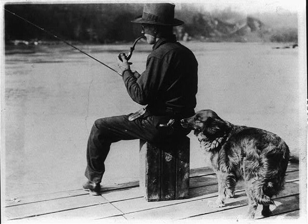 Vintage man fishing off dock with dog and smoking pipe.