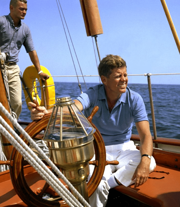 Jfk john f kennedy on a sailboat smiling in wind with a man standing behind him. 