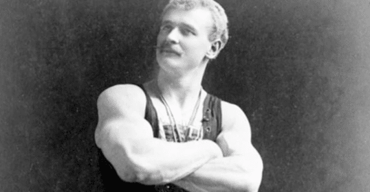 A vintage black and white photo of a mustachioed strongman posing bigger and stronger with his arms crossed.