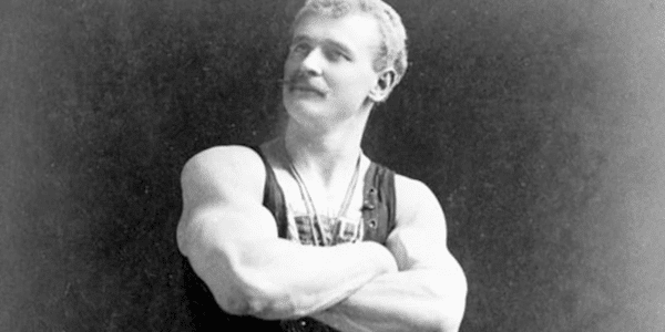 A vintage black and white photo of a mustachioed strongman posing bigger and stronger with his arms crossed.