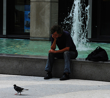 A man sitting on a ledge next to a fountain, deep in thought, as a pigeon peacefully coexists nearby.