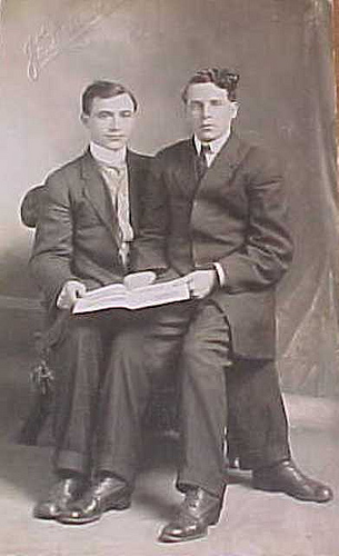 Vintage male friend sitting on thigh of another while wearing suite and holding book in hands.